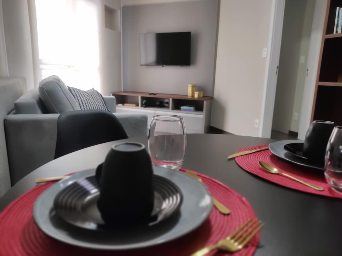 A dining table is set with black plates and cups, featuring gold utensils and a glass. In the background, a small living area includes a gray sofa and a wall-mounted television, creating a functional and welcoming space.