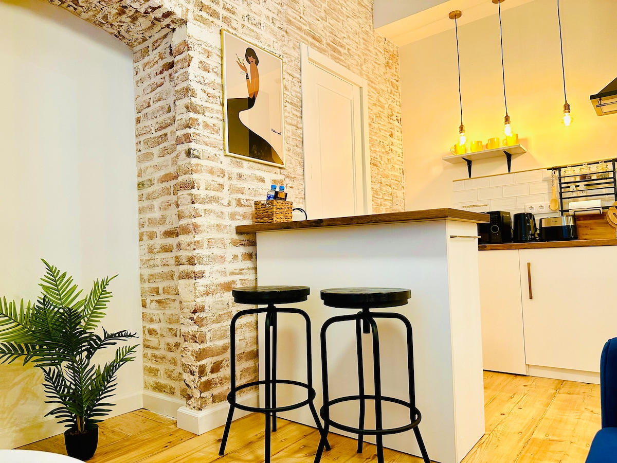 A sleek kitchen area features a wooden countertop with two black bar stools positioned alongside. The open space is highlighted by exposed brick walls and modern lighting fixtures. A small potted plant adds a touch of greenery, enhancing the kitchen's overall appeal.