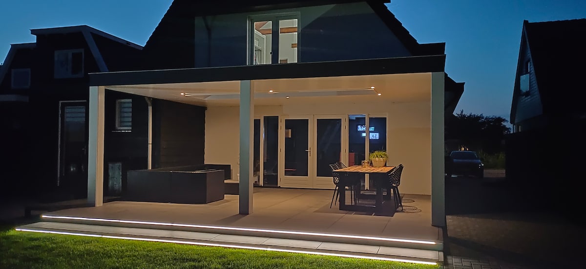 The covered outdoor patio is illuminated softly at night, showcasing a dining table with chairs and a seating area. A jacuzzi is positioned nearby, enhancing the tranquil ambiance. Large glass doors lead into the interior, and the exterior features a clean, modern aesthetic.