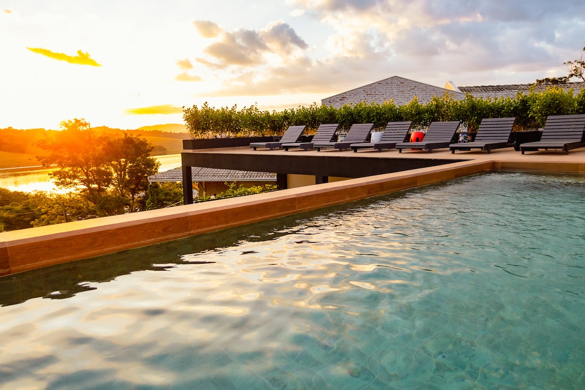 A tranquil pool area is captured at sunset, with several lounge chairs arranged beside it. The water reflects the warm light, while lush greenery borders the space, enhancing the natural setting. In the background, a serene landscape features trees and the glimmer of water.