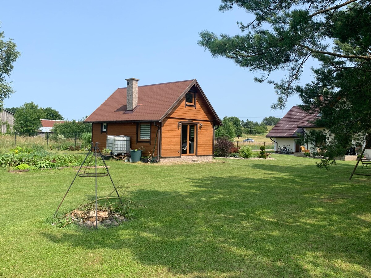 A charming wooden house is set against a clear blue sky, surrounded by a well-maintained lawn. A fire pit is positioned in the foreground, with a tripod grill frame. The adjacent fields add to the serene countryside atmosphere.