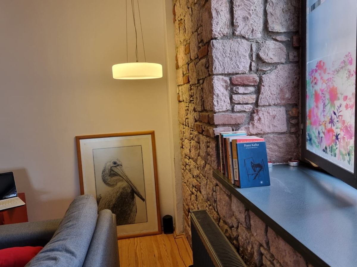A cozy corner features a soft grey sofa beside a window with colorful floral designs. A framed artwork of a pelican leans against the wall, next to a small stack of books. Exposed stone walls add texture to the indoor space.