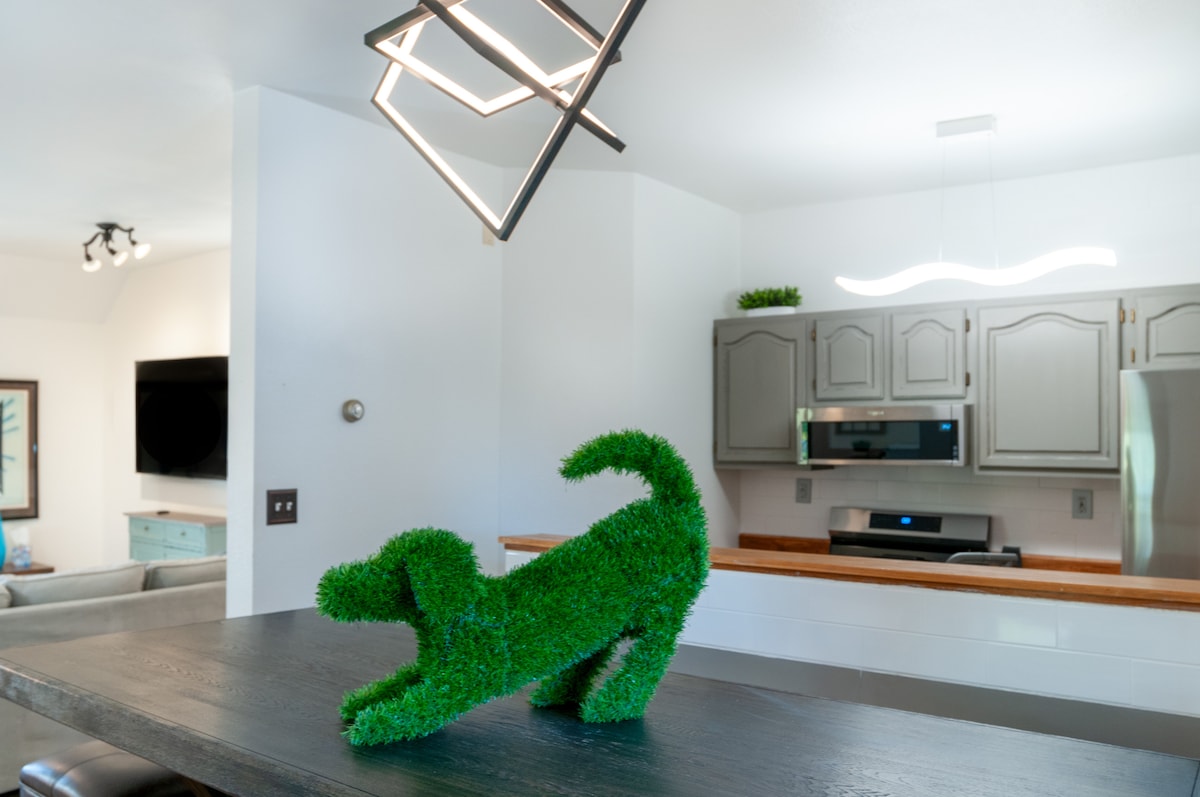 A unique green dog sculpture made of artificial grass is positioned on a dark wooden countertop. The background reveals a modern kitchen with light-colored cabinetry and stainless steel appliances, illuminated by contemporary lighting fixtures above.