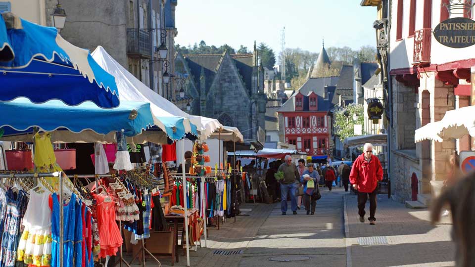 A lively market street features colorful tents and stalls displaying various clothing and goods. Visitors stroll along the paved path, with historic buildings visible in the background. The scene captures a blend of local culture and community engagement in a charming town setting.