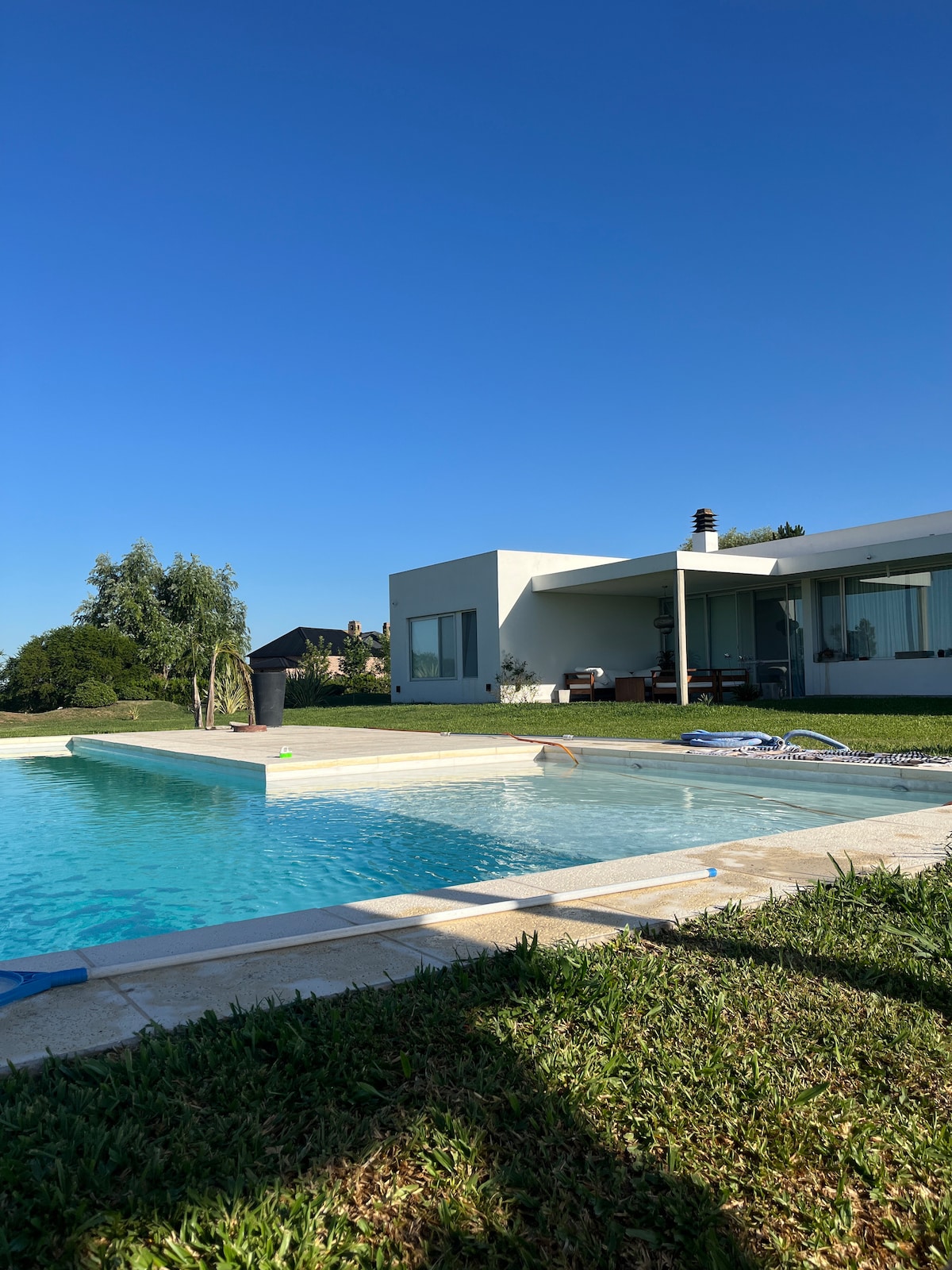 A modern house with large glass windows is visible beside a clear swimming pool. The outdoor area features a spacious lawn with trees, and a patio seating area is seen in the background. A blue sky provides a bright backdrop to the scene.