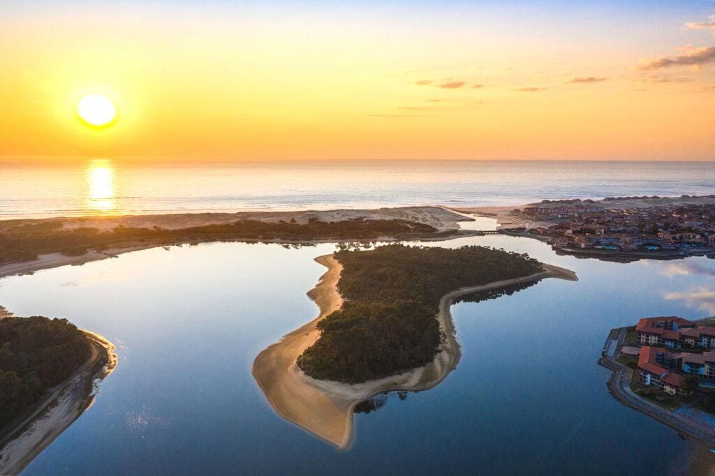 A serene aerial view captures the sun setting over a calm body of water, reflecting pastel hues of orange and pink. An island covered with greenery emerges from the water, while sandy shorelines frame the landscape, blending the natural surroundings with distant coastal developments.