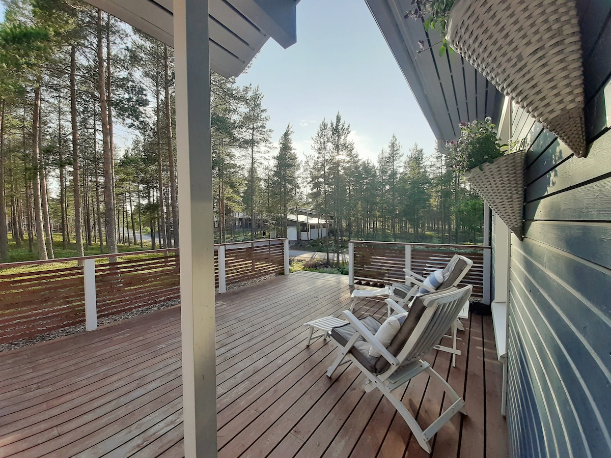 A spacious wooden terrace is featured, surrounded by tall trees that provide a sense of tranquility. Two loungers are positioned for relaxation, and decorative planters hang from the exterior wall, adding a touch of nature to the setting.