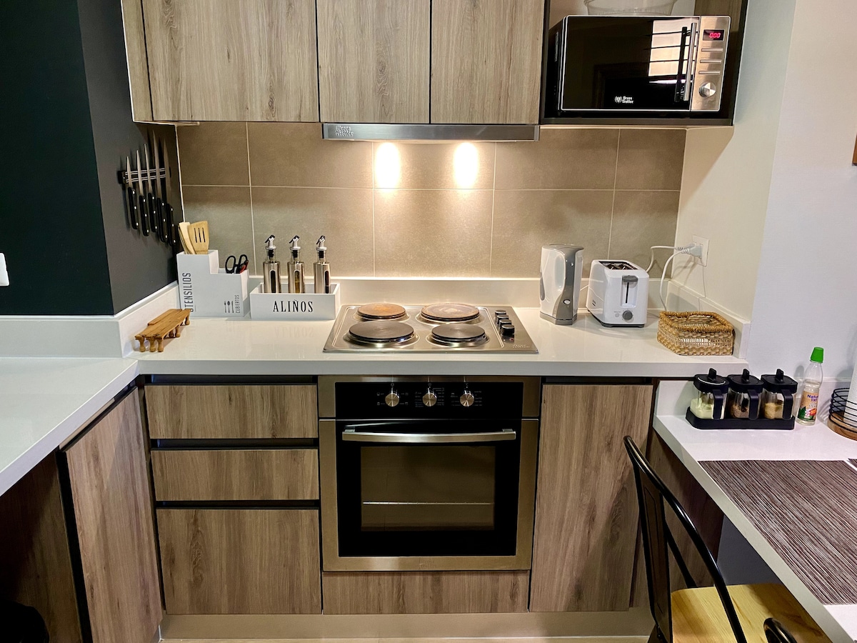 A well-equipped kitchen features modern wooden cabinetry and a clean countertop. Cooking appliances, including a stovetop and an oven, are prominently displayed. Small appliances like a microwave and kettle are also present, along with essential utensils and condiments arranged neatly on the countertop.