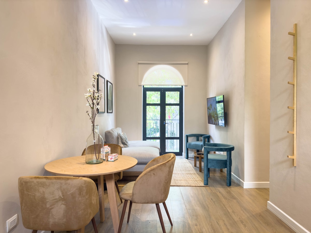 A modern living space features a round wooden dining table surrounded by two upholstered chairs. A light-colored sofa is positioned along one wall, complemented by a flat-screen TV mounted nearby. Natural light enters through a large window with a sliding door opening to a balcony.