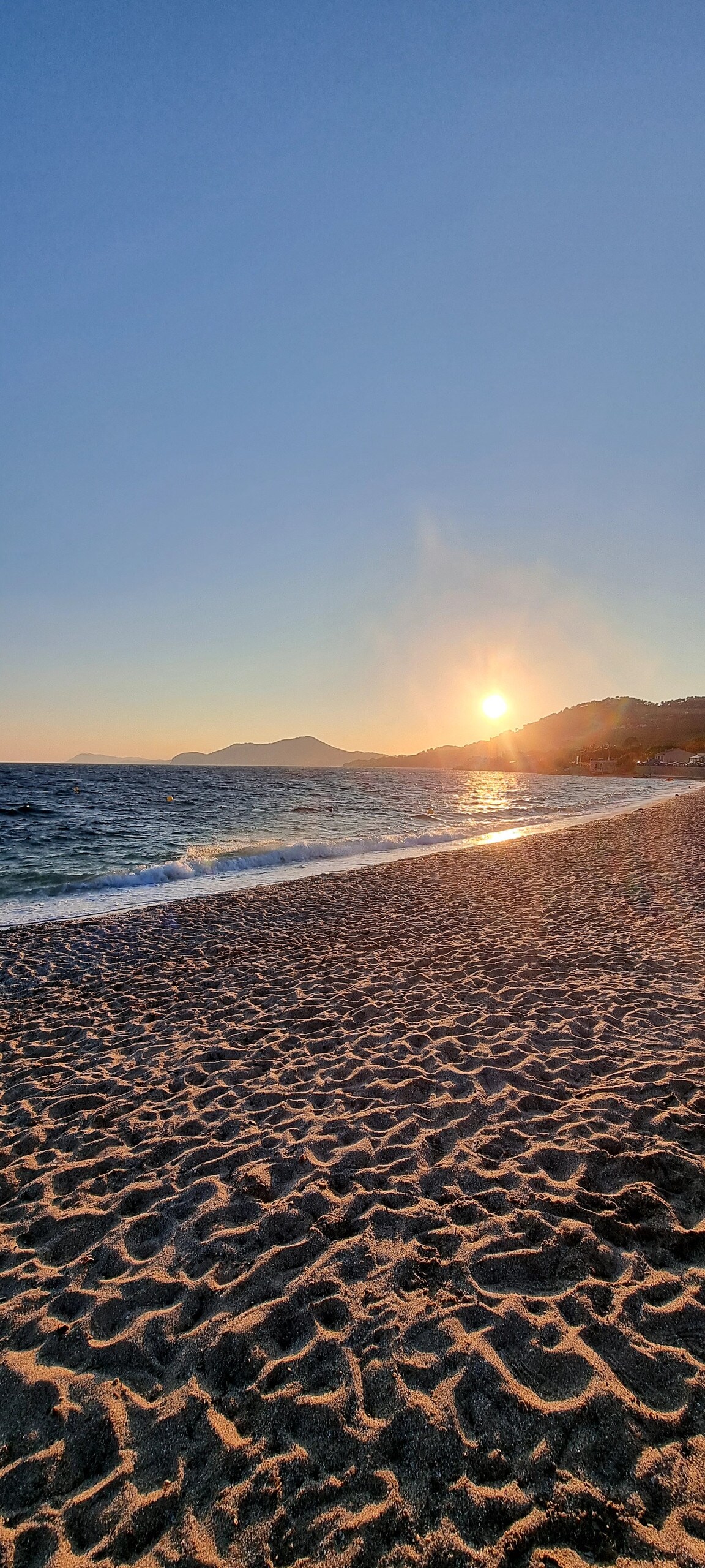 A serene beach scene captures the sun setting over gentle waves. The golden light reflects off the water, while soft sand stretches along the shoreline. Silhouetted mountains form a backdrop against the vibrant sky, offering a sense of tranquility and natural beauty.