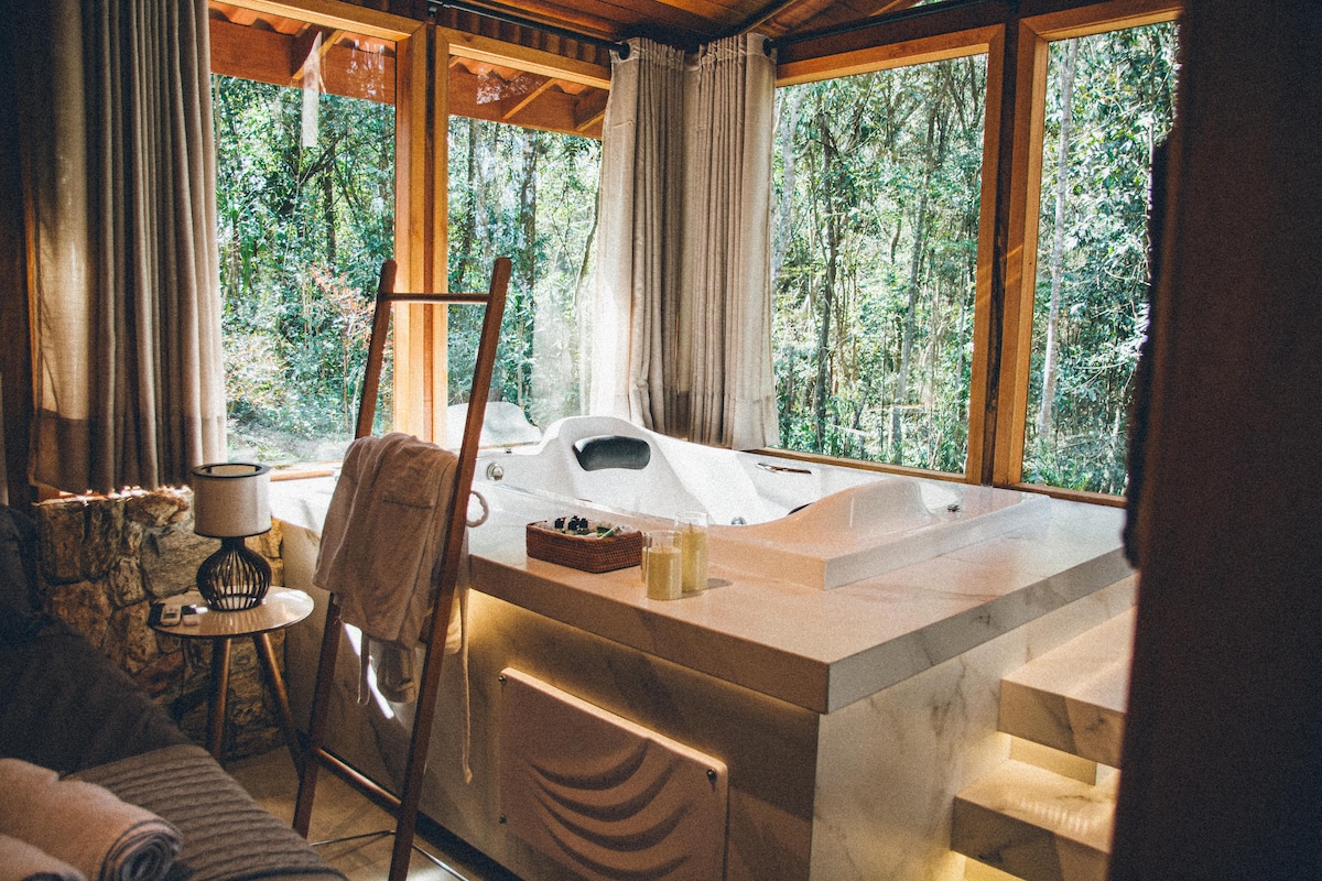 A spacious hot tub is positioned beside large windows, allowing natural light to fill the area. Surrounding greenery is visible through the glass, enhancing the serene environment. Soft towels are draped over a nearby ladder, and minimalist decor complements the relaxing space.