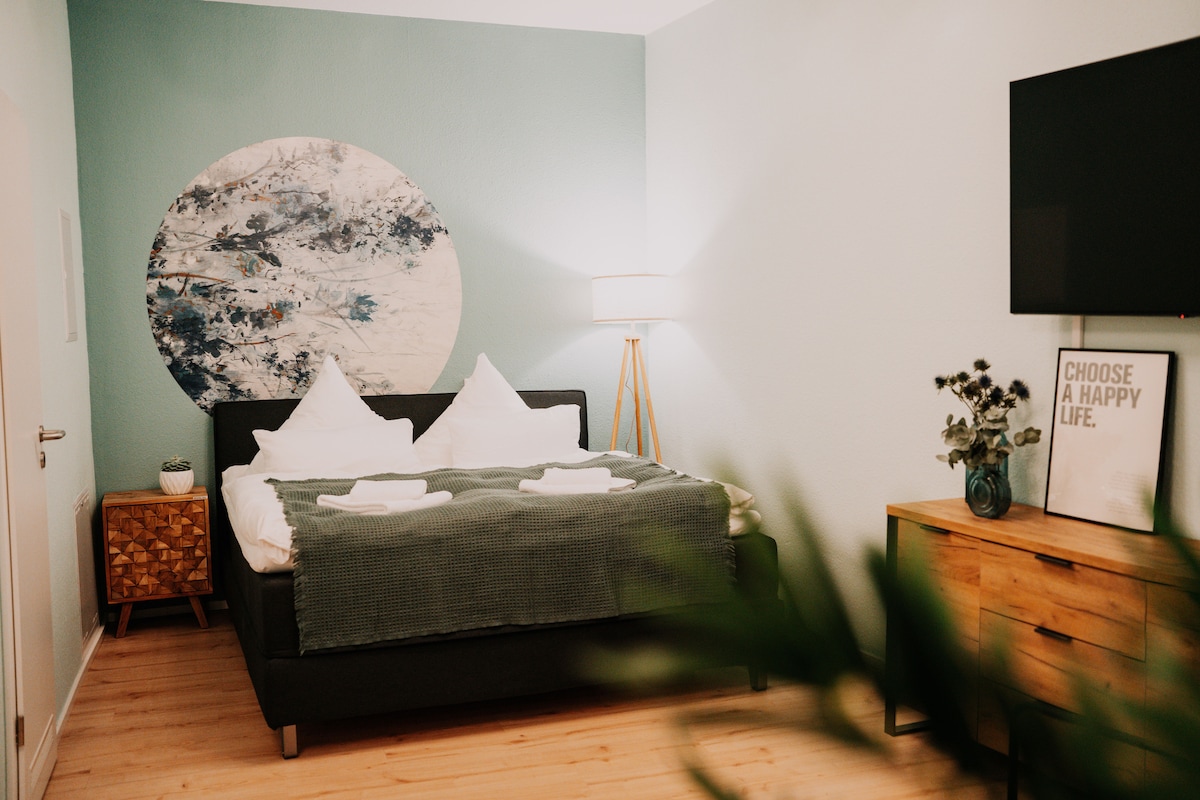 A serene bedroom is depicted, featuring a king-size bed with soft pillows and a cozy green blanket. A light-colored wooden dresser and a modern TV are positioned on the opposite wall. Natural light illuminates the room, complemented by a stylish lamp beside the bed.