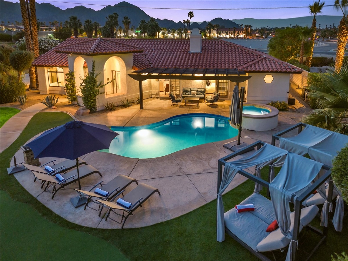 A tranquil outdoor space is showcased with a swimming pool and spa, surrounded by lounge chairs and an umbrella. Two cabanas offer shaded seating. Palm trees line the area with mountain views in the background, creating a serene atmosphere for relaxation.