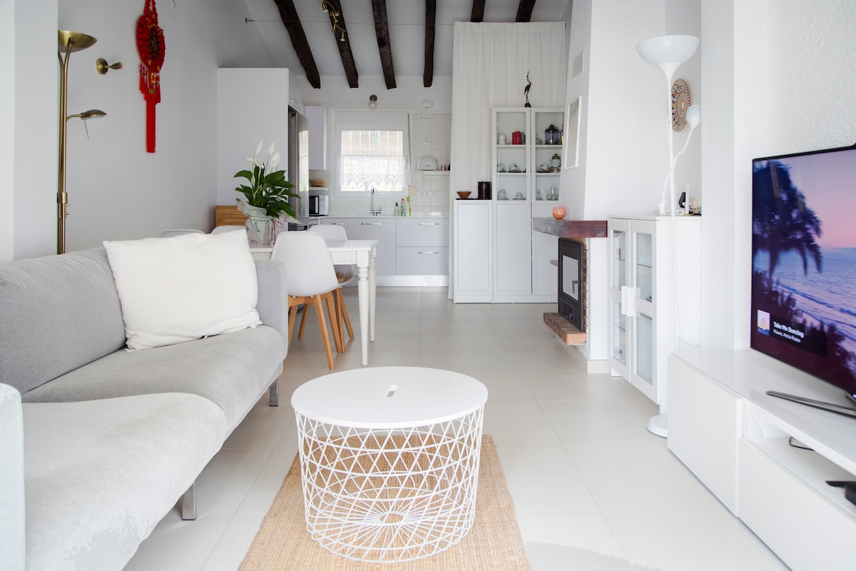 A spacious living area features a light-colored sofa adorned with soft pillows. A round coffee table sits atop a natural fiber rug. In the background, a dining space with a white table and chairs is visible, alongside a modern kitchen equipped with white cabinetry.