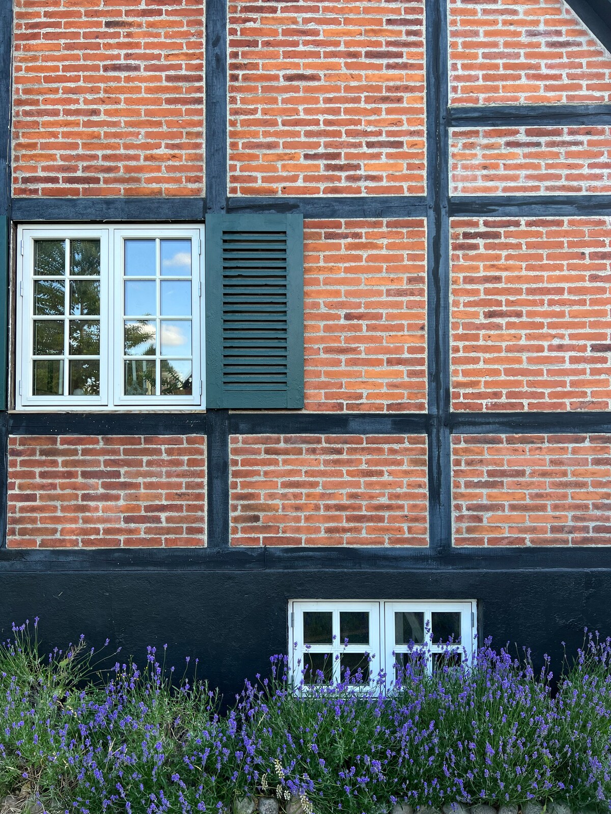 The exterior of the house features a classic brick facade with black timber framing. Two windows are positioned above a smaller window, adorned by a blooming lavender garden below. Traditional green shutters provide a charming accent to the overall rustic appearance.