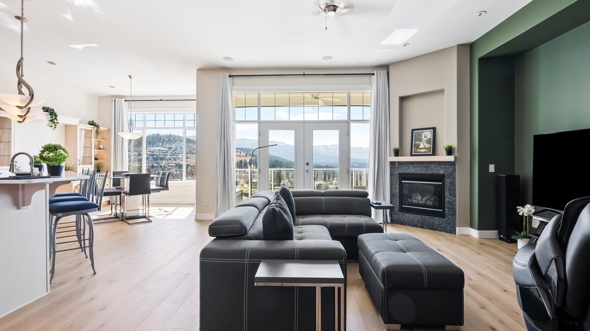 A spacious living area is presented with a modular black sectional sofa and an ottoman, complemented by large windows offering expansive views of the surrounding landscape. A sleek entertainment unit and a dining area with stylish chairs are also visible, creating a modern, inviting atmosphere.