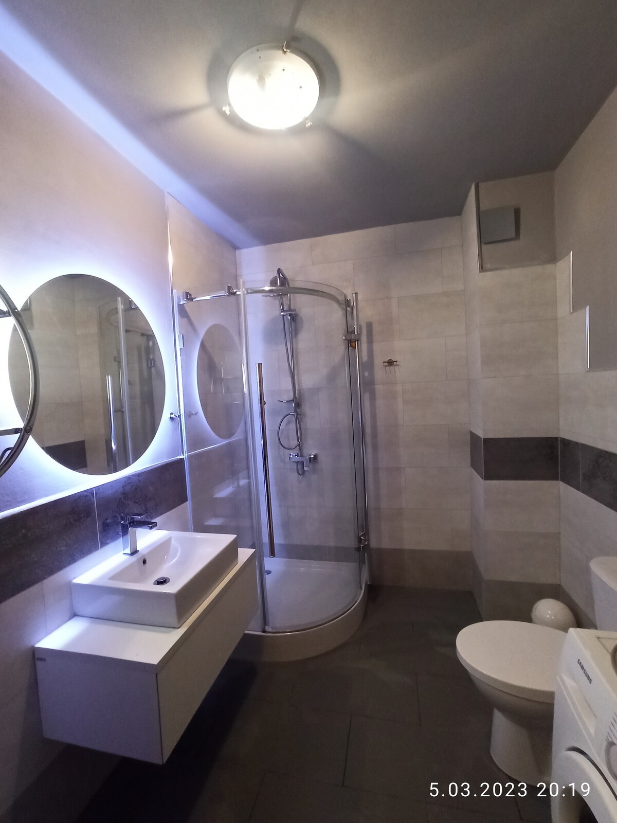 The bathroom is designed with modern fixtures, featuring a walk-in shower with clear glass, a compact sink with a mirror above, and a toilet. Soft lighting is provided by an overhead fixture, and the tiled walls are complemented by dark accents.