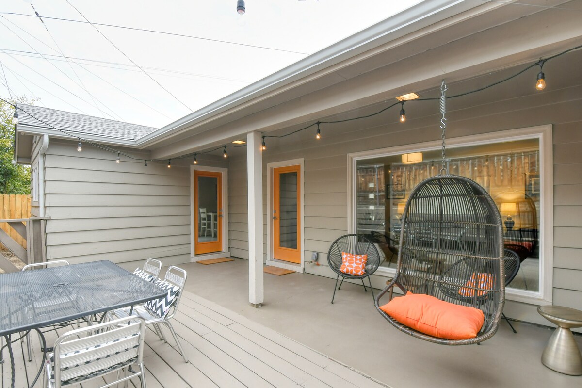 A spacious outdoor patio area features a hanging swing chair with bright orange cushions alongside a dining table with multiple metal chairs. Soft lighting is provided by decorative string lights overhead, creating a casual atmosphere suitable for relaxation and dining.