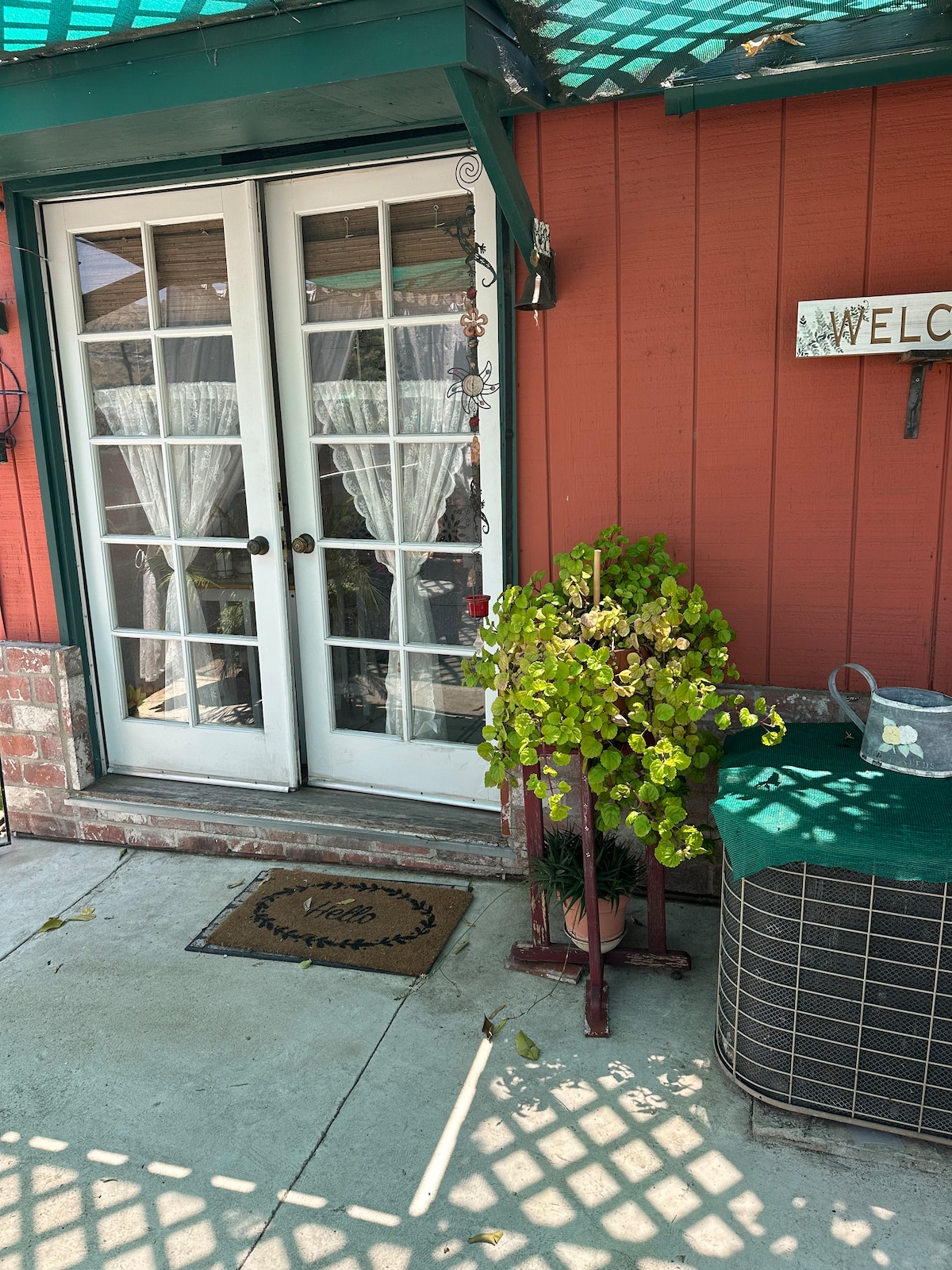 Double doors with glass panels are featured in a vibrant red exterior. A welcome mat is placed on a concrete pathway, alongside a potted plant and a small table covered with a green cloth. Natural light filters through sheer curtains behind the doors.