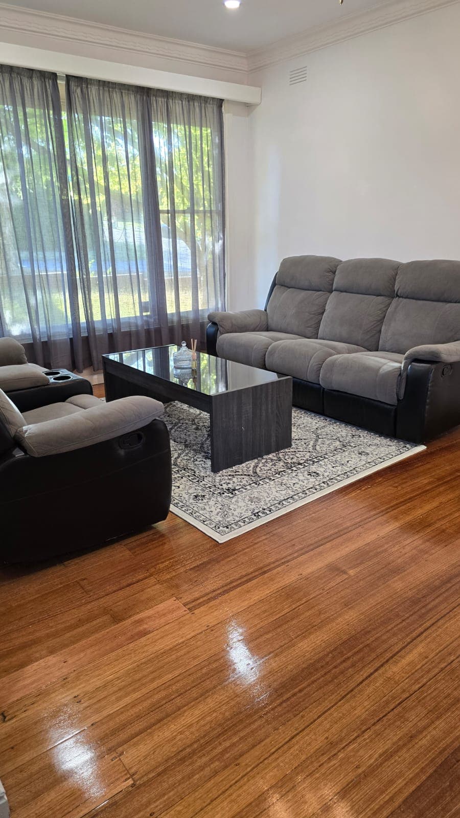 A living area features a comfortable gray sofa and matching armchairs positioned around a sleek glass coffee table. A patterned rug sits beneath the table, complementing the polished wooden floor. Floor-to-ceiling windows allow natural light to illuminate the space.