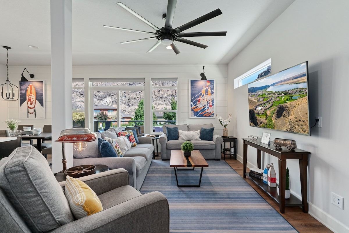 A bright living area features a comfortable seating arrangement with gray sofas and a wooden coffee table. Large windows provide views of the outdoors, while a smart TV and ceiling fan enhance the space. Artwork adds a colorful touch to the walls.