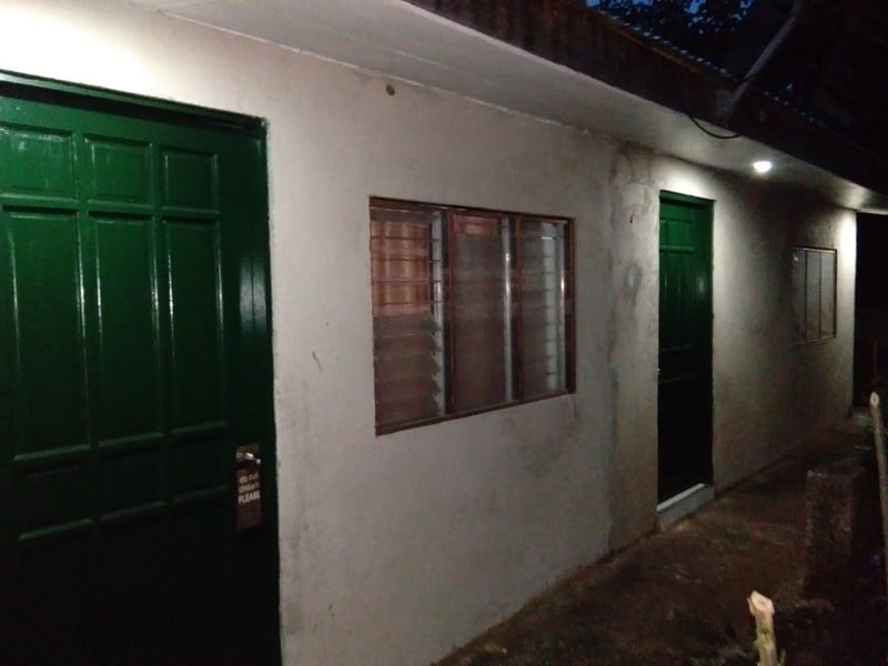 The exterior of a simple building is shown, featuring two green doors and a window with horizontal slats. Soft lighting highlights the entrance area, while the walls are painted in a neutral tone, contributing to the overall functional and welcoming appearance.