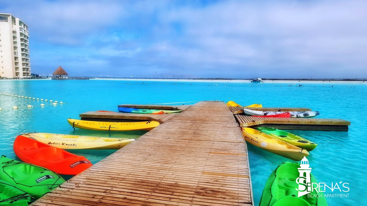 A wooden dock extends over clear turquoise waters, lined with various brightly colored kayaks. The calm surface reflects the overcast sky, while a distant shoreline is visible in the background. The scene conveys a serene atmosphere ideal for water activities.
