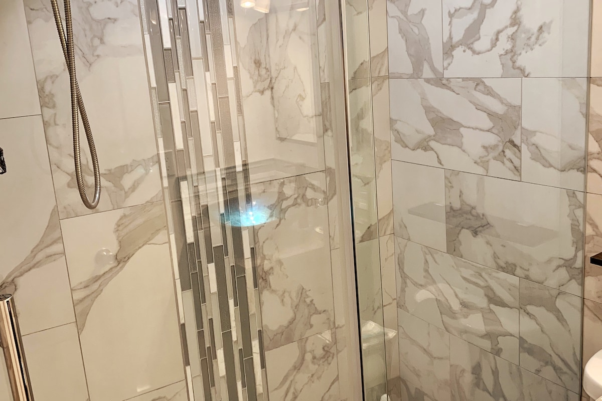 A modern shower area is showcased with sleek glass doors and marble-patterned tiles. The space is well-lit, featuring a built-in shelf for toiletries. A rainfall showerhead is positioned above, creating a clean and refreshing appearance.