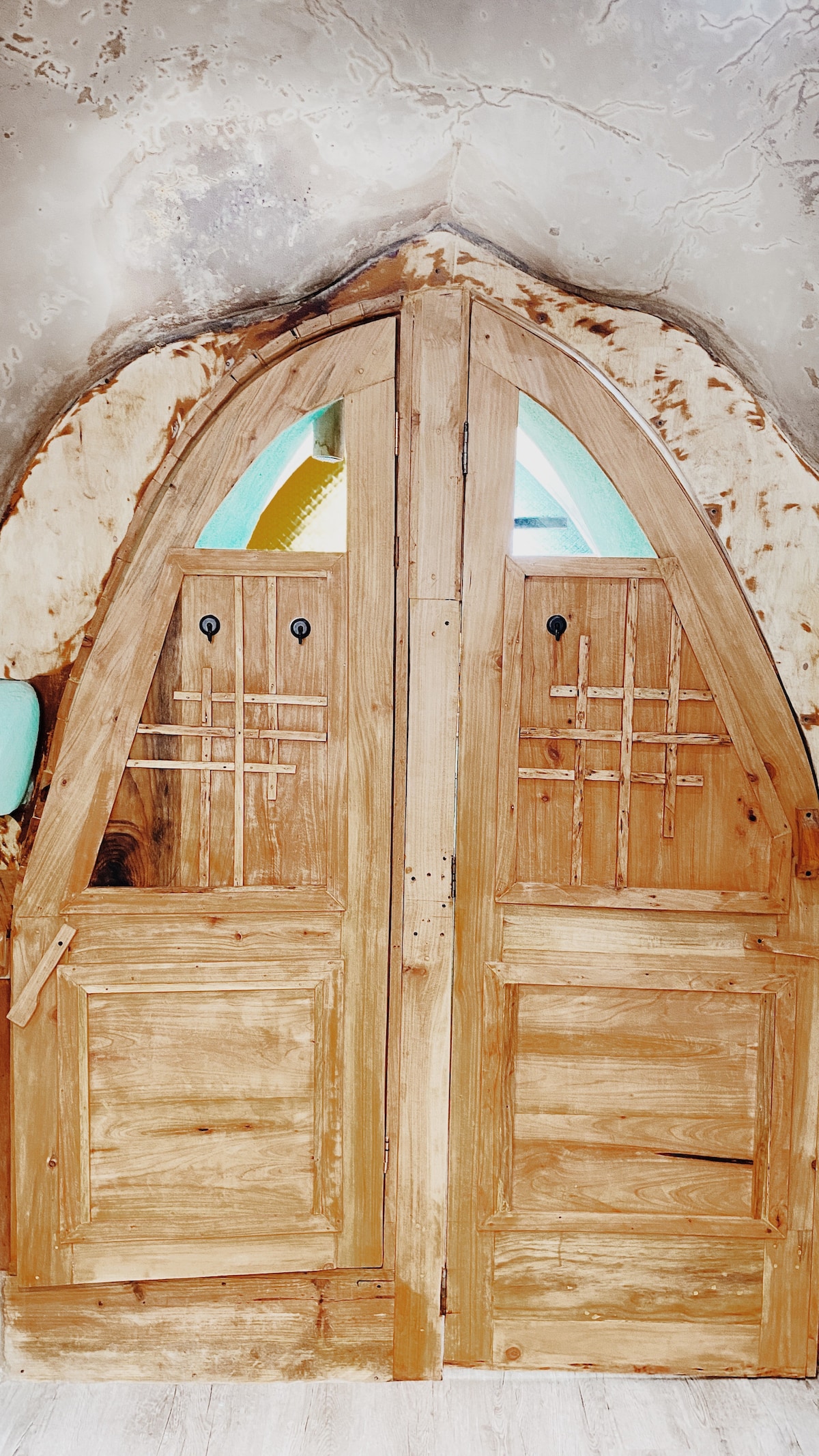 A rustic wooden double door features an intricate design with diagonal carvings. The door's natural wood grain and finish highlight its craftsmanship, while round door handles add a touch of detail. The arch at the top complements the overall organic appearance of the entrance.