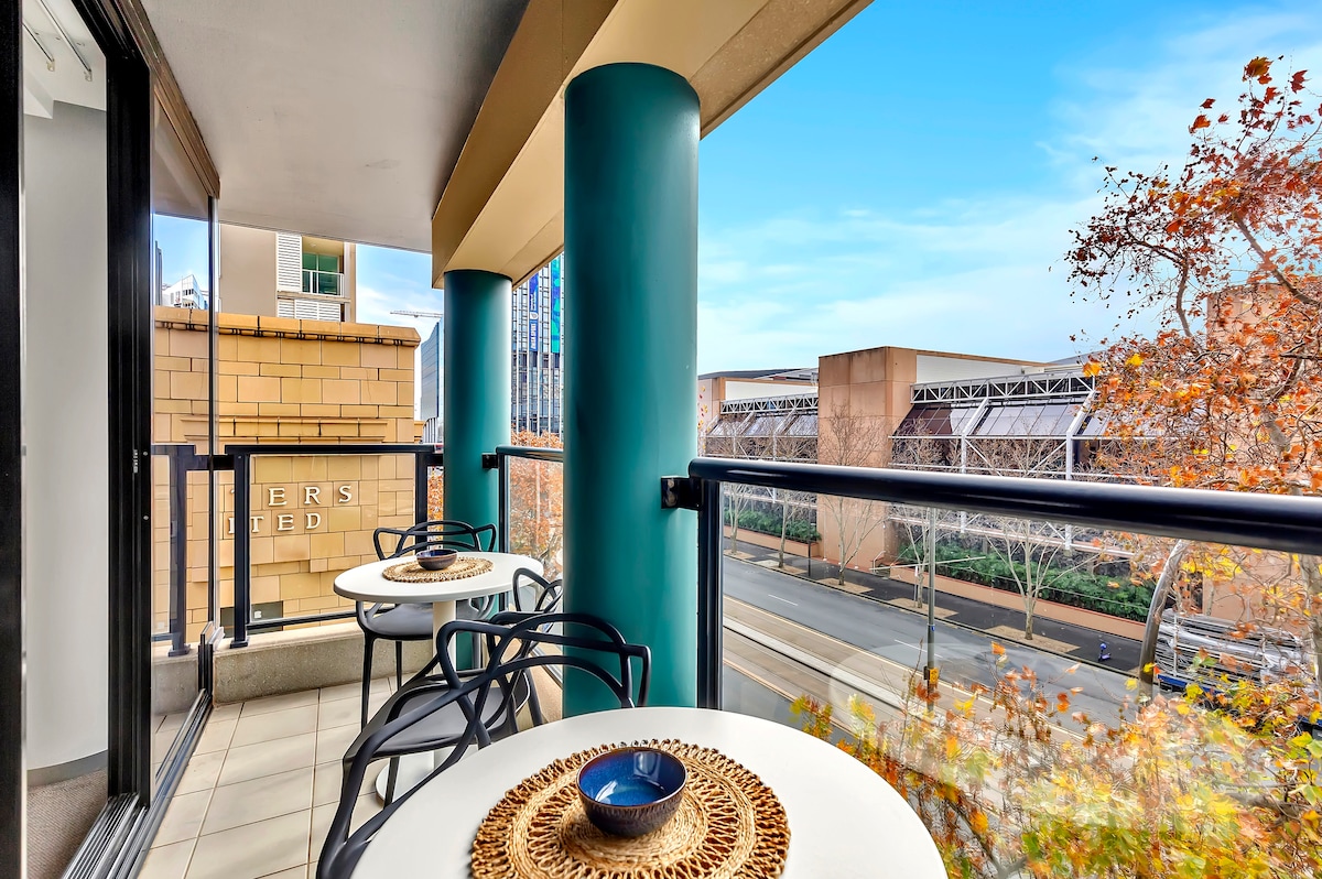 The balcony offers dual two-seater table settings with woven placemats, overlooking a tree-lined street. Pillars in a calming hue support the overhang, while a clear sky is visible. The outdoor area provides a serene space for dining or enjoying fresh air.