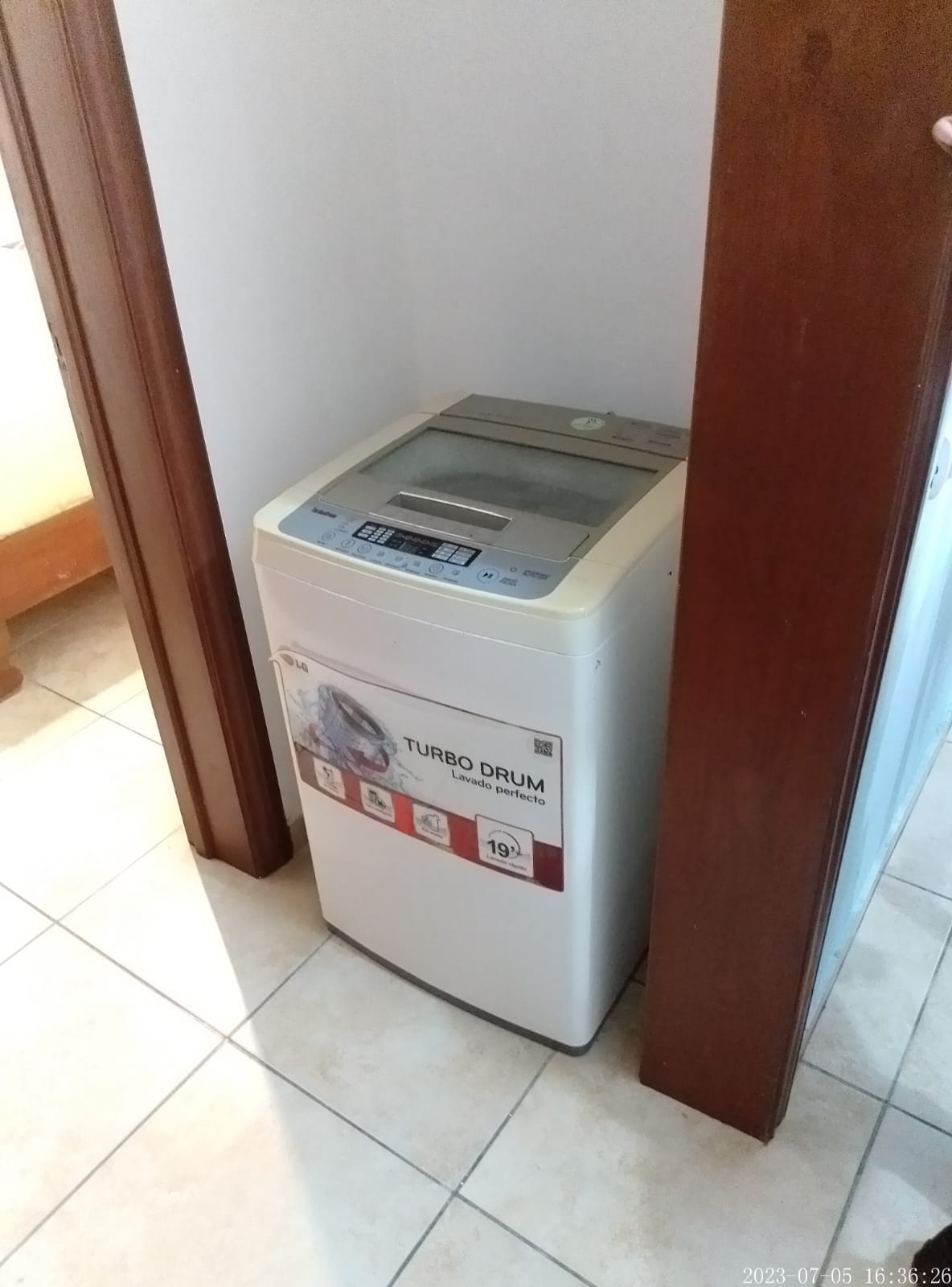 A washing machine is positioned between two doorways, featuring a top-loading design. The machine displays a control panel on its lid, which is marked with brand and model information. Neutral tile flooring surrounds the appliance, adding to the utility space's simplicity.