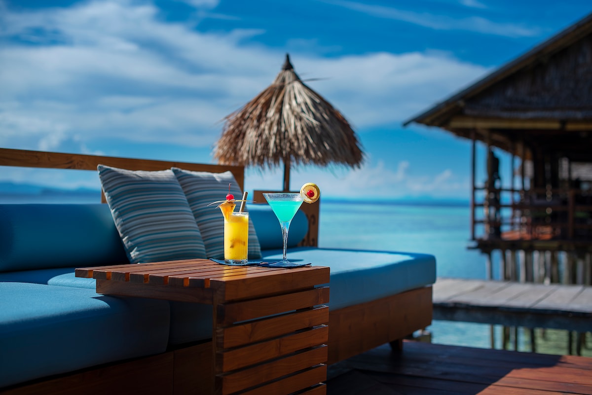 A cozy seating area features a wooden table adorned with two colorful cocktails, one garnished with a slice of fruit. A shaded over-water bungalow is visible in the background, with clear blue water and a bright sky enhancing the tranquil setting.