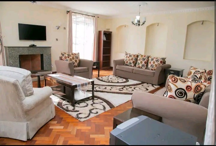 A spacious living area features soft seating options arranged around a central coffee table, set on a decorative rug. Natural light filters through large windows, illuminating the room's warm tones and contrasting patterns on the cushions. A television and bookshelf are positioned against the wall.