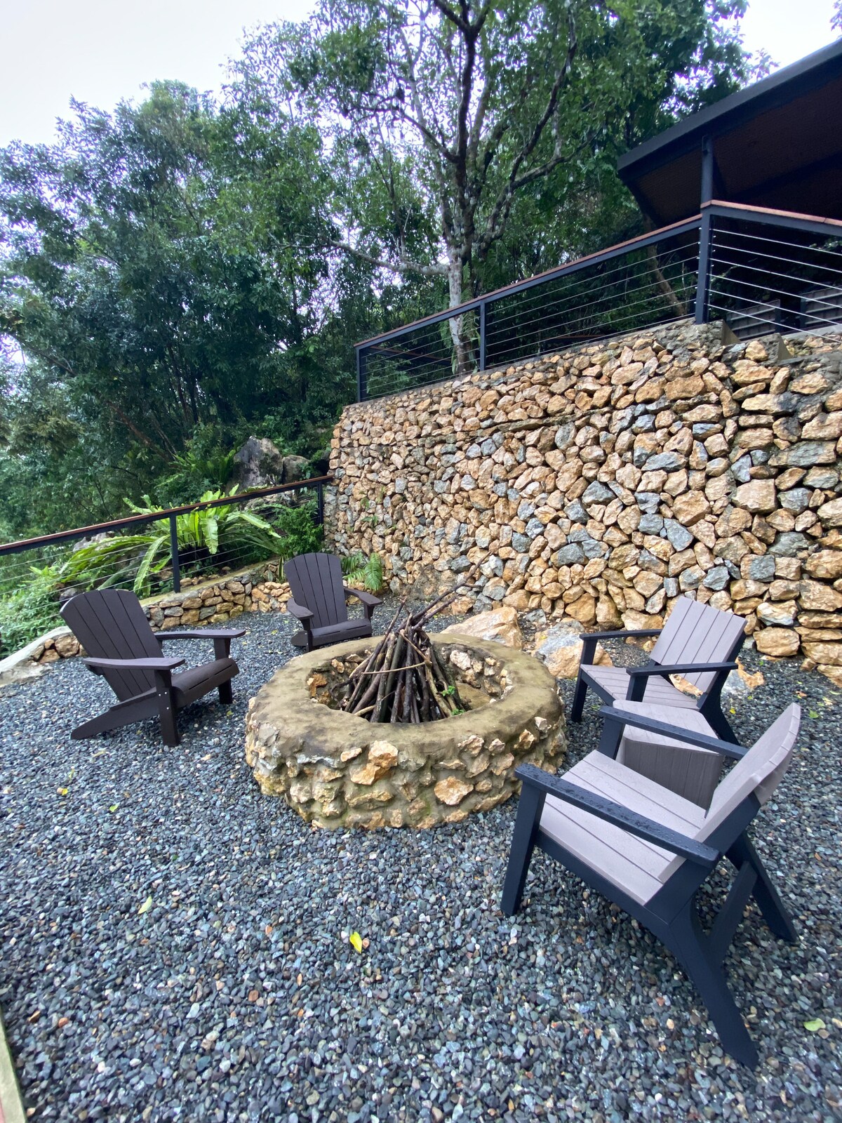 A fire pit surrounded by smooth stones is set in a gravel area, accompanied by four comfortable wooden chairs. Lush greenery is visible in the background, with a stone wall rising beside the structure, contributing to the serene outdoor setting.