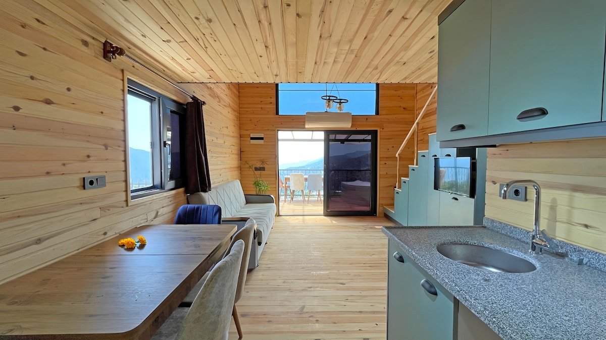 The interior features a light wood paneling that creates a warm atmosphere. A dining table with chairs is positioned next to a cozy seating area, while a well-equipped kitchen is visible. Large windows provide a view of the expansive landscape beyond.