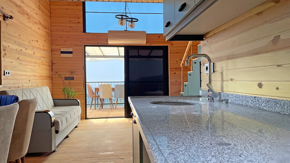 The interior space features a modern kitchen with a grey countertop and a sink. A cozy sofa is positioned against a wooden wall. Glass doors lead to an outdoor dining area, allowing natural light to fill the room while providing pleasing views of the landscape.