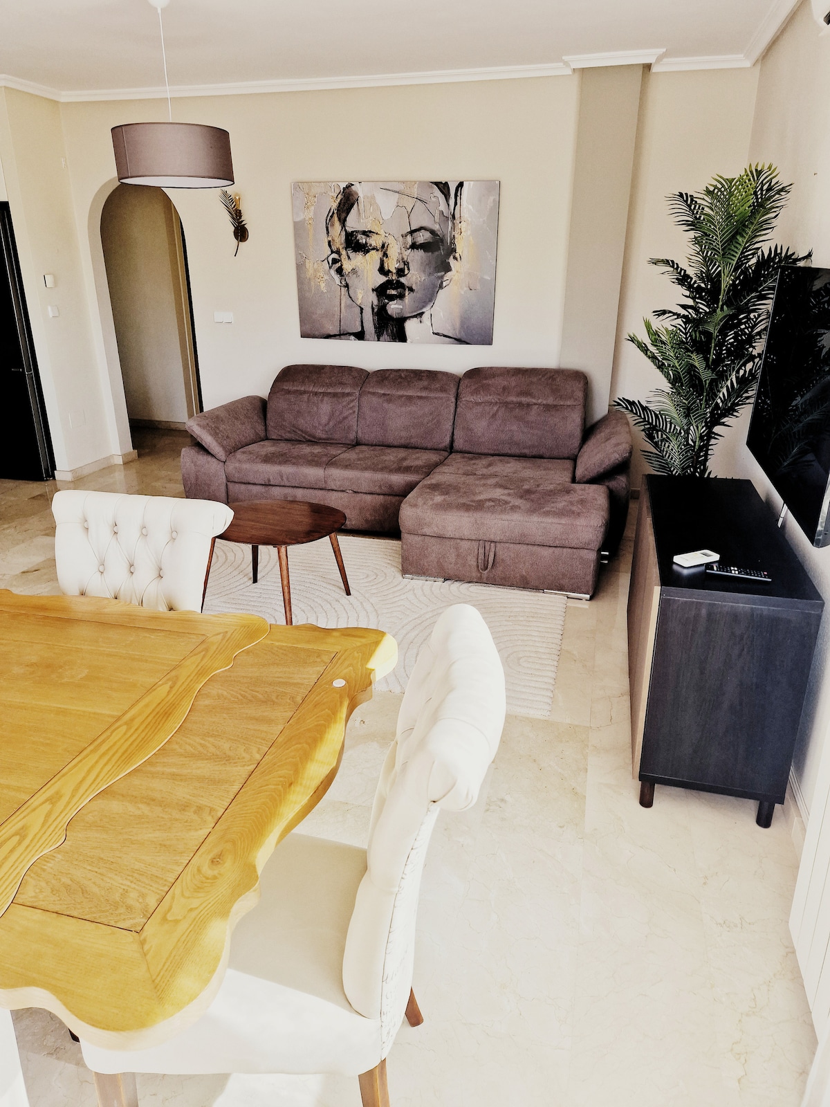 A spacious living and dining area is depicted, featuring a comfortable brown sofa and a wooden dining table surrounded by upholstered chairs. A television is placed on a dark media console, complemented by decorative plants and soft lighting from an overhead fixture.