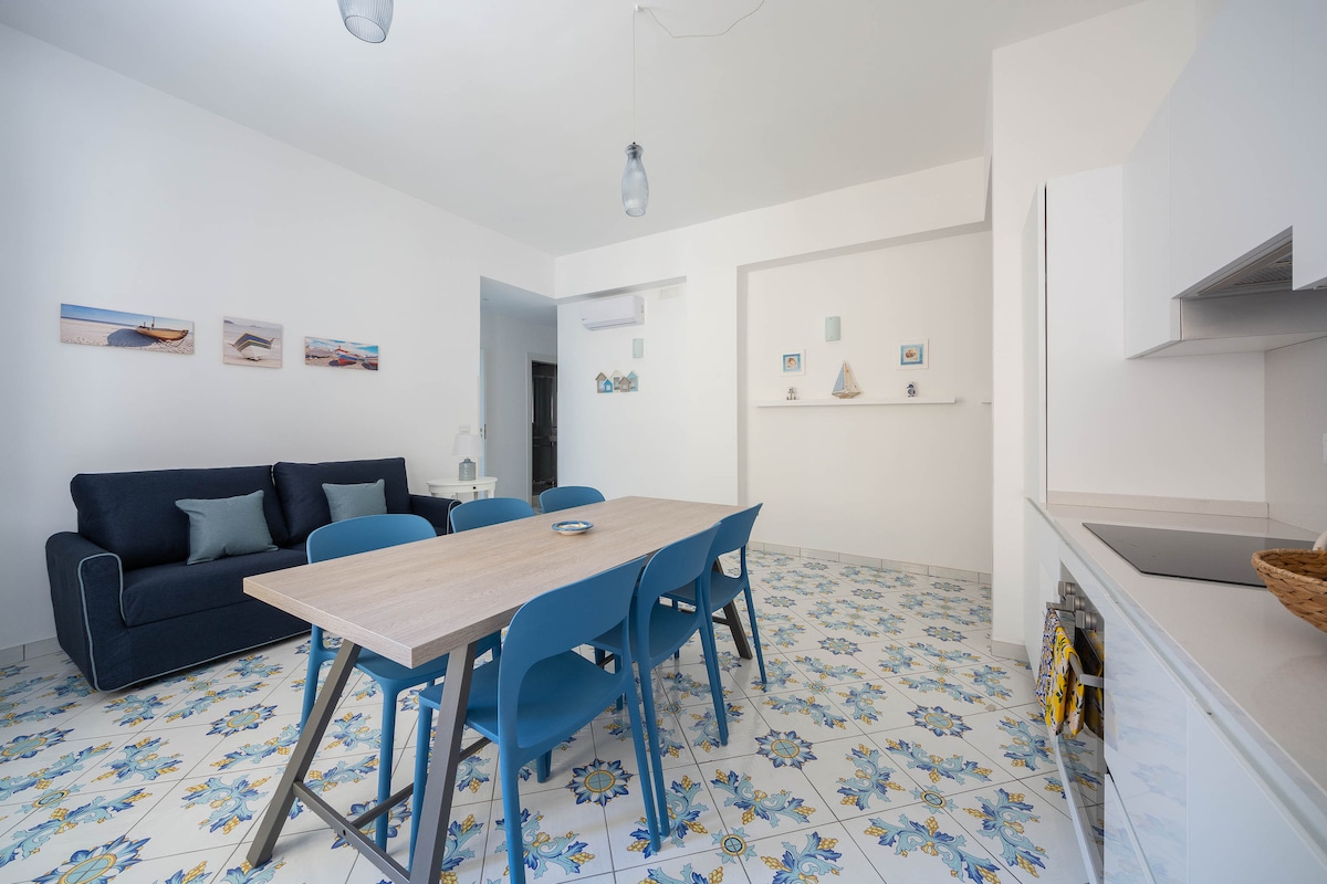 A bright and spacious living/dining area is depicted with modern furnishings. A large wooden dining table accommodates six blue chairs. A comfortable navy sofa is positioned nearby. The floor features colorful majolica tiles, enhancing the room's light atmosphere, while subtle artwork adorns the walls.