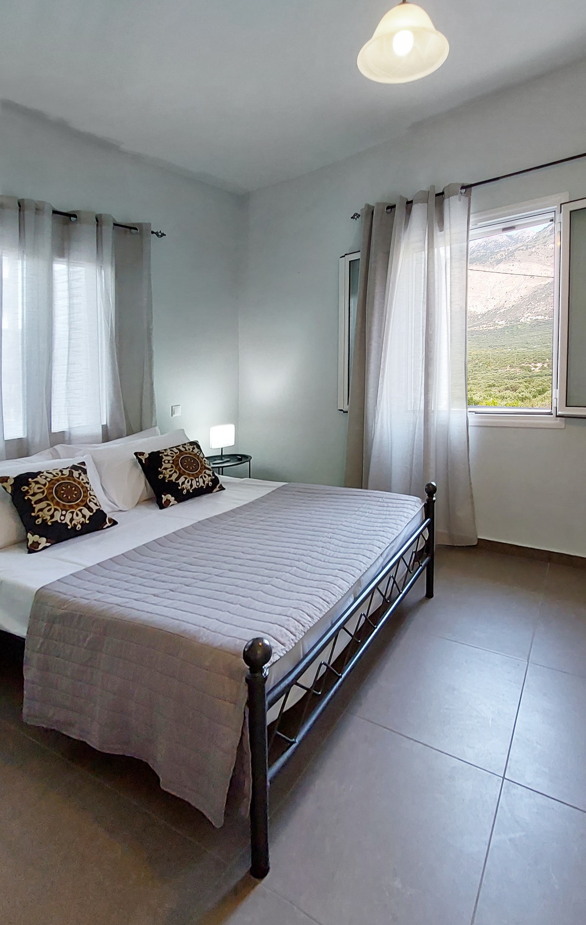 A bright bedroom features a spacious bed adorned with decorative pillows and a light grey blanket. Large windows allow natural light to fill the room, while soft curtains frame the view of the surrounding landscape. A simple bedside table and lamp complete the inviting atmosphere.
