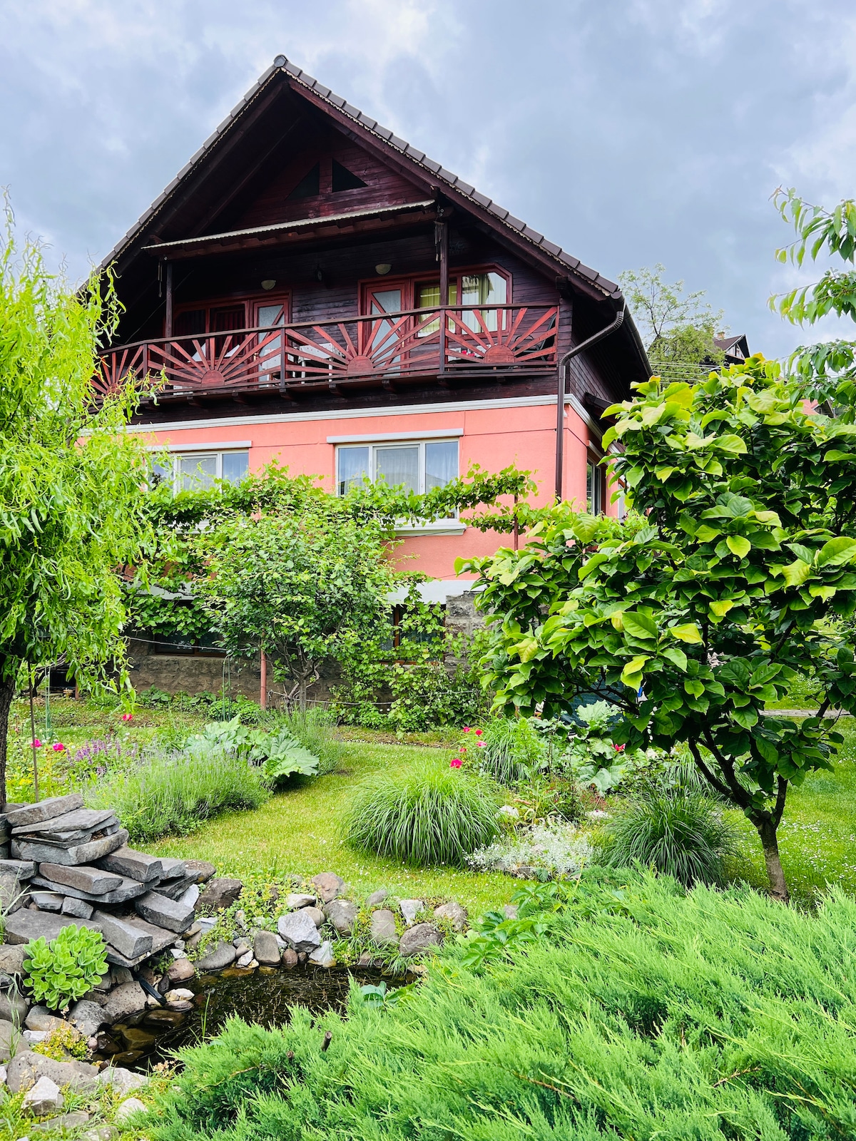 A charming three-story building is surrounded by a lush garden. The exterior features a combination of wooden balconies and pink walls, with greenery and colorful flowers enhancing the serene atmosphere. A small pond is partially visible among the well-maintained plants in the foreground.