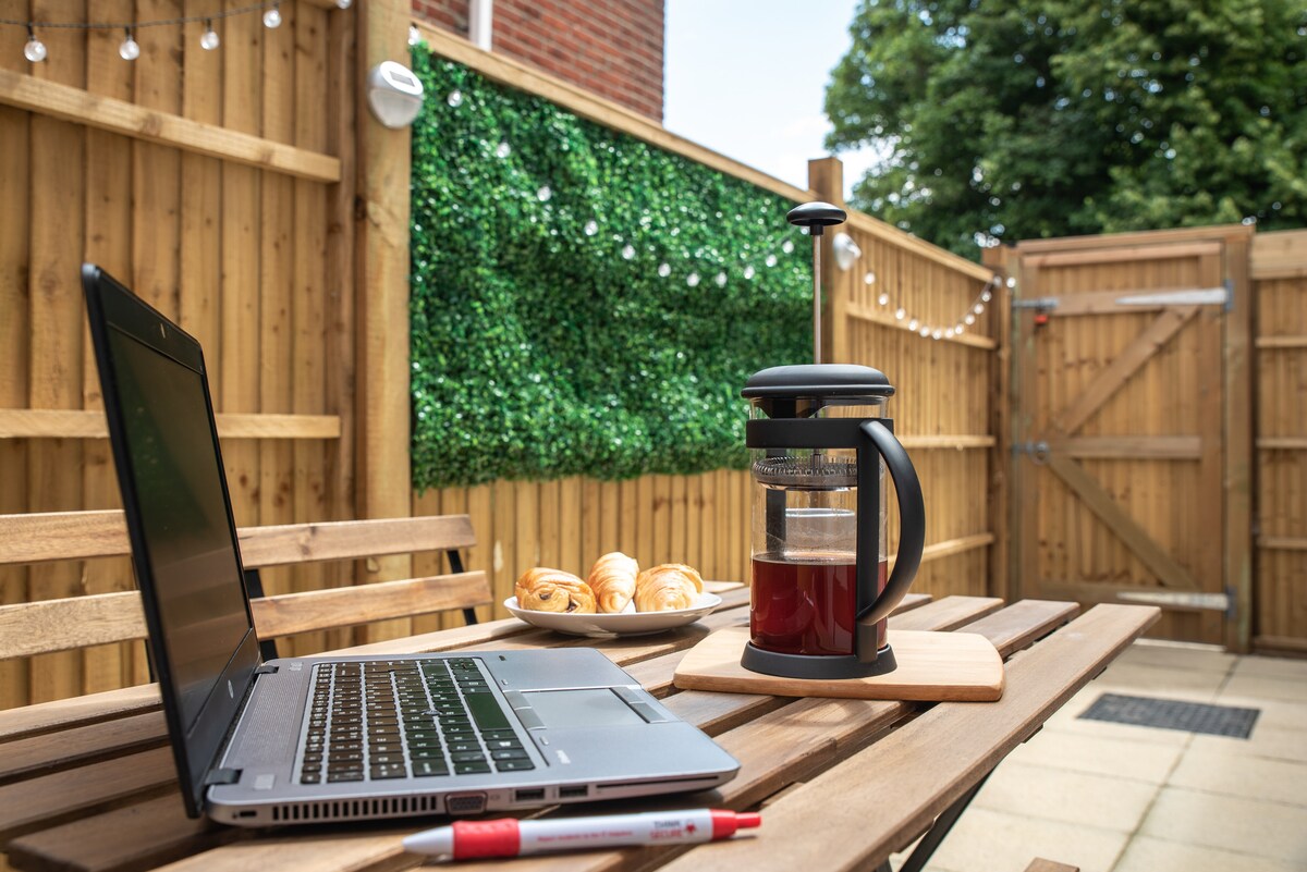 An outdoor courtyard area features a wooden table with a laptop and a French press coffee maker. A wooden serving board holds freshly baked pastries. The backdrop is adorned with green foliage and a wooden fence, providing a cozy and private atmosphere.