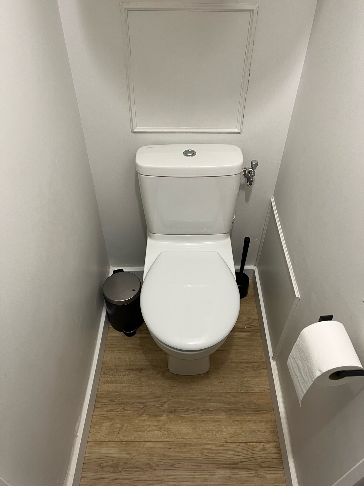 A clean and minimalistic bathroom offers a white toilet, complemented by a toilet paper holder and a waste bin nearby. Light wooden flooring enhances the spacious feeling of this well-maintained area.