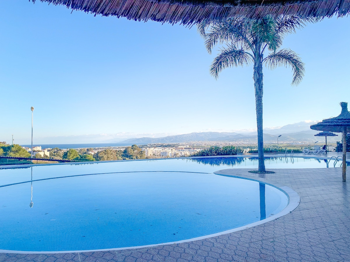 An inviting infinity pool reflects the clear sky, surrounded by well-maintained landscaping. A palm tree casts a gentle shadow over the pool area, offering a serene view of the horizon where the land meets the sea.