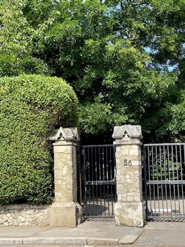 A private entrance gate is framed by lush greenery and mature trees, creating a sense of seclusion. The gate features iron bars and stone pillars, with the number '56' clearly displayed on one pillar, indicating the entrance to the Vicarage.