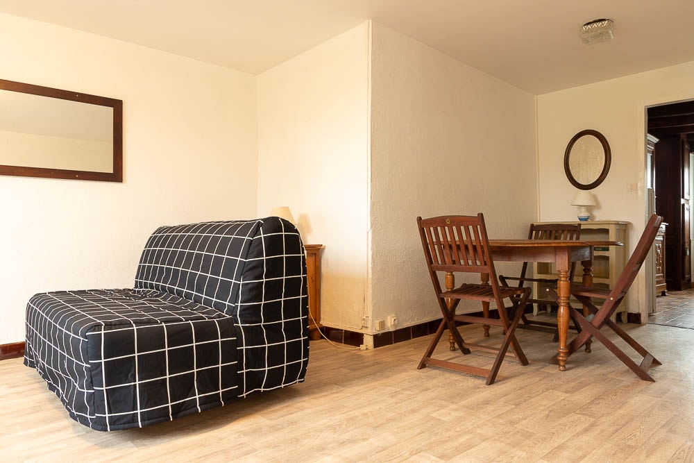 A bright living area features a convertible sofa covered in a grid-patterned upholstery. A round wooden dining table is surrounded by folding chairs. Natural light illuminates the space, creating a relaxed atmosphere, while wall mirrors add depth to the room.