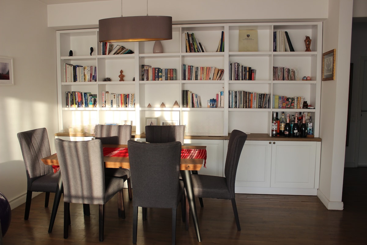 A dining area is composed of a wooden table surrounded by six upholstered chairs. A well-organized shelf displays a collection of books and decorative items. Natural light filters through a nearby window, illuminating the warm, wooden floor.