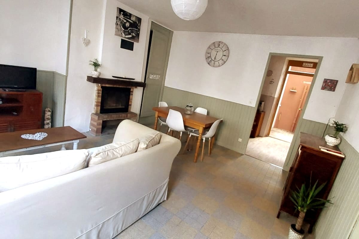 A living area features a comfortable sofa facing a wall-mounted TV. A wooden coffee table sits in front, while a dining table with four white chairs occupies the corner. A brick fireplace adds a cozy touch, and a wall clock is displayed above.