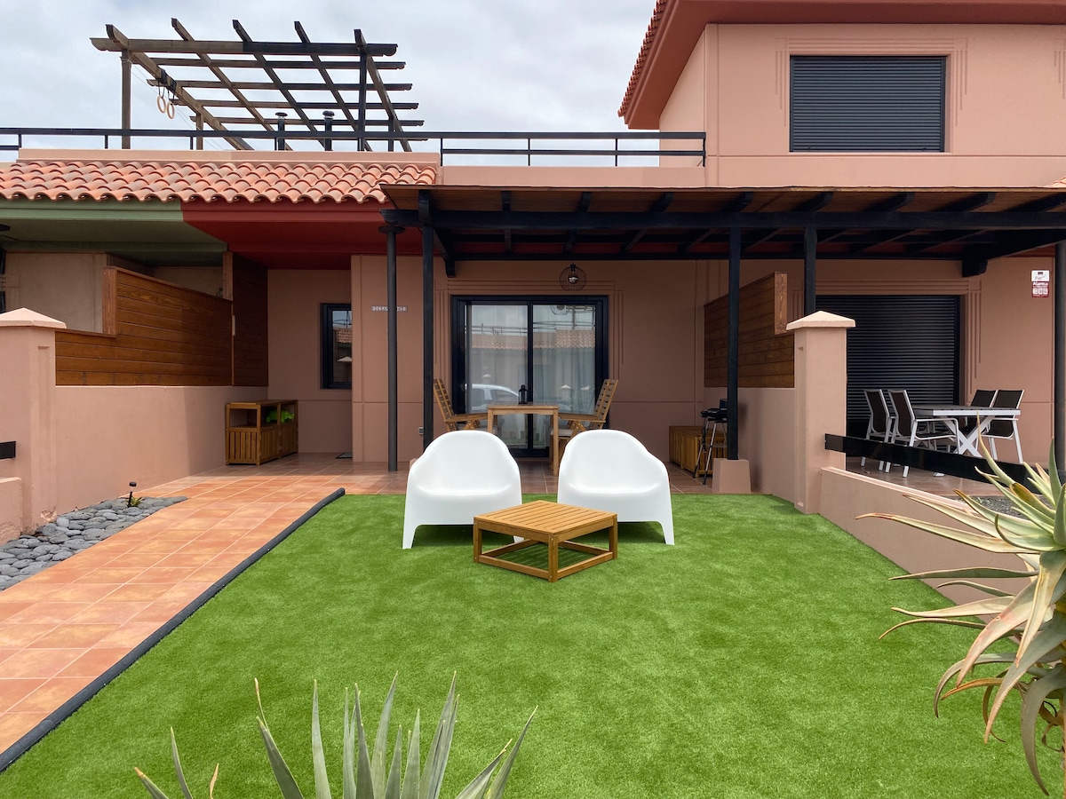 A serene outdoor space features artificial grass with two chill-out chairs positioned beside a wooden table. The area is framed by a covered terrace, showcasing a mix of lounge and dining seating options, complemented by stone pathways and surrounding greenery.