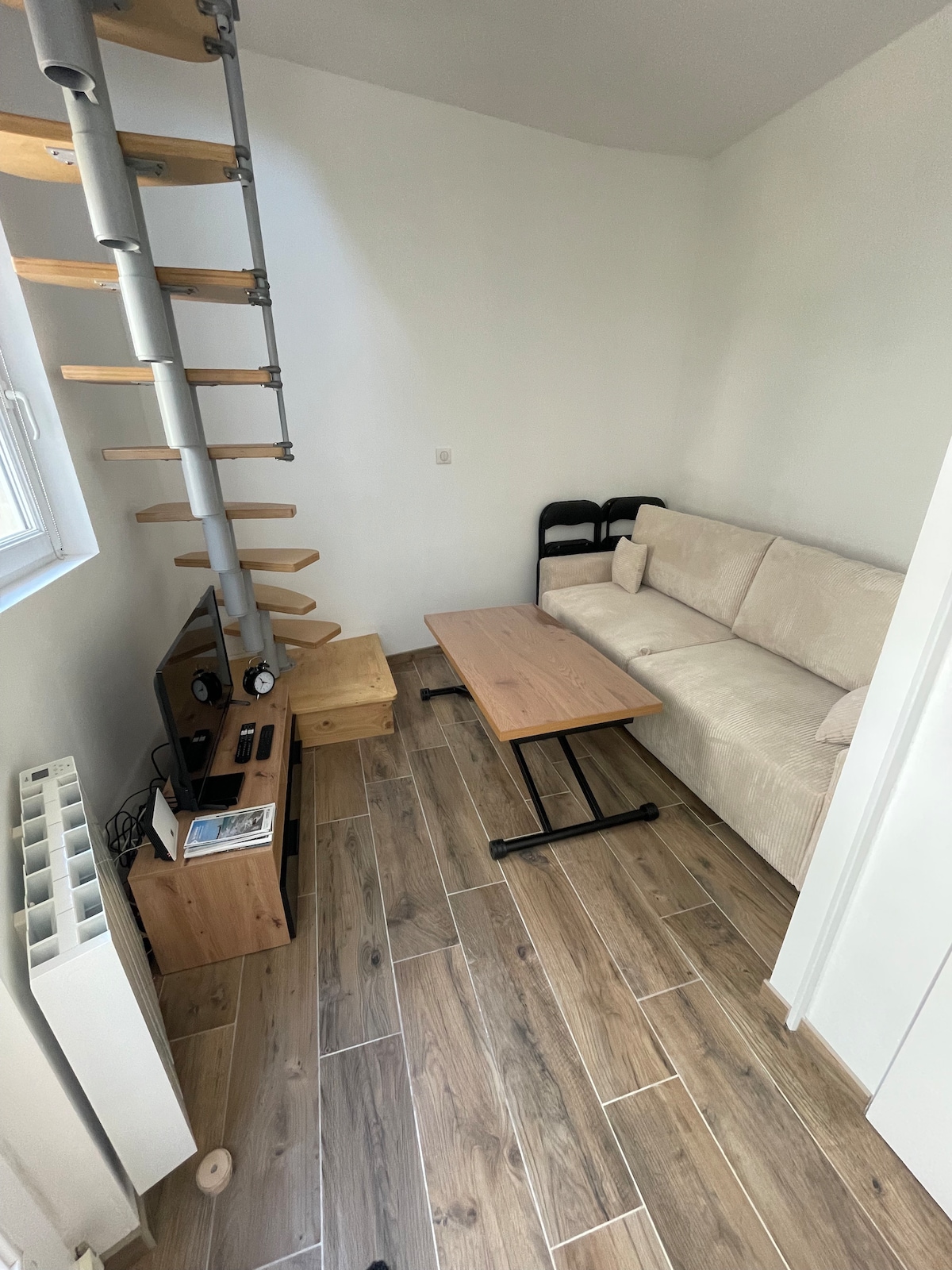 A compact living area features a soft, light-colored sectional sofa facing a wooden coffee table. A wall-mounted TV and media console are positioned nearby. Light-colored, wooden flooring complements the overall design, while a spiral staircase leads to the upper level.
