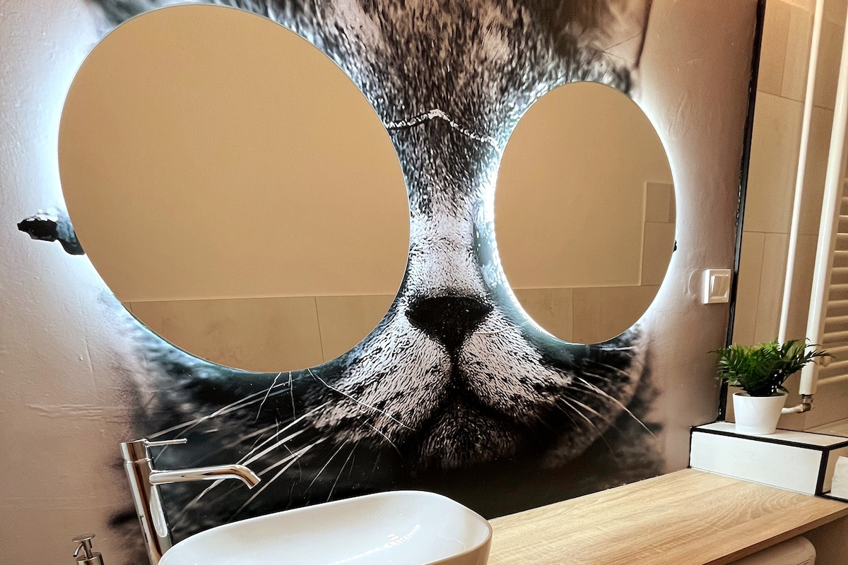 A unique bathroom features a large, artistic mirror shaped like cat glasses, illuminated along the edges. Below, a modern sink with a sleek faucet is placed on a wooden countertop. A small plant adds a touch of greenery to the space, enhancing its charm.
